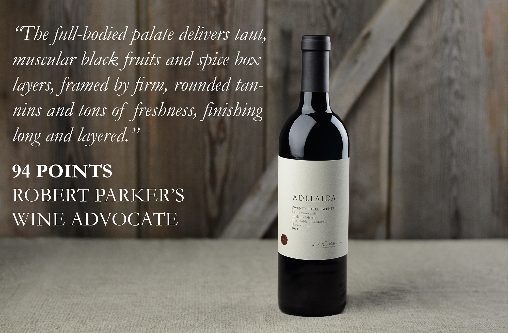 Bottle of Twenty Three Twenty on a table with a wood background. Text says: “The full-bodied palate delivers taut, muscular black fruits and spice box layers, framed by firm, rounded tannins and tons of freshness, finishing long and layered. 94 points Robert Parker’s Wine Advocate 